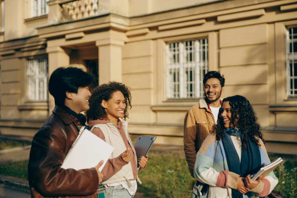 Grupo de quatro estudantes sorrindo e conversando em frente a um prédio universitário, em clima descontraído e ensolarado.