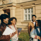 Grupo de quatro estudantes sorrindo e conversando em frente a um prédio universitário, em clima descontraído e ensolarado.