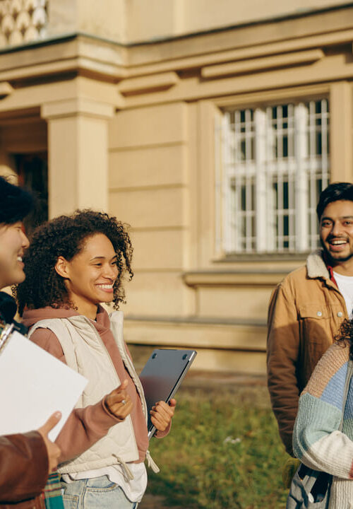 Grupo de quatro estudantes sorrindo e conversando em frente a um prédio universitário, em clima descontraído e ensolarado.