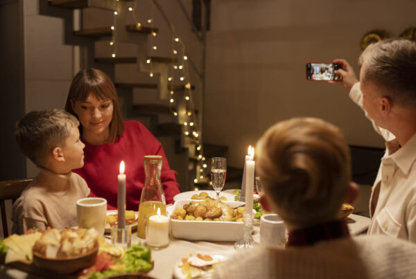 A imagem mostra uma cena de jantar em família durante o Dia de Ação de Graças. Uma mulher de blusa vermelha e uma criança estão sentados próximos, interagindo de maneira carinhosa. A mesa está posta com pratos típicos, incluindo um peru assado, saladas e bebidas. Em segundo plano, uma escada decorada com luzes pisca-pisca adiciona um toque festivo ao ambiente, enquanto uma pessoa ao fundo tira uma foto do momento, capturando a celebração familiar.