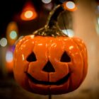 A imagem mostra uma típica abóbora de Halloween, esculpida com um rosto sorridente, com olhos e boca triangulares. A abóbora, de cor laranja, está destacada em primeiro plano, com um fundo desfocado que exibe luzes coloridas e suaves, sugerindo uma decoração ou ambiente festivo. A iluminação da cena dá um tom acolhedor e ao mesmo tempo misterioso, comum nas celebrações de Halloween.