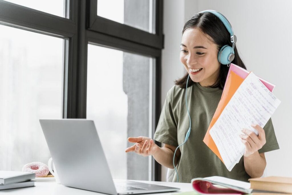 A imagem mostra uma jovem participando de uma videoconferência em um ambiente de estudo. Ela está sorrindo, usando fones de ouvido e olhando para um laptop à sua frente. A jovem segura alguns papéis e uma pasta colorida, sugerindo que está envolvida em uma aula online ou reunião de trabalho. A cena é iluminada por uma grande janela ao fundo, criando uma atmosfera clara e acolhedora.