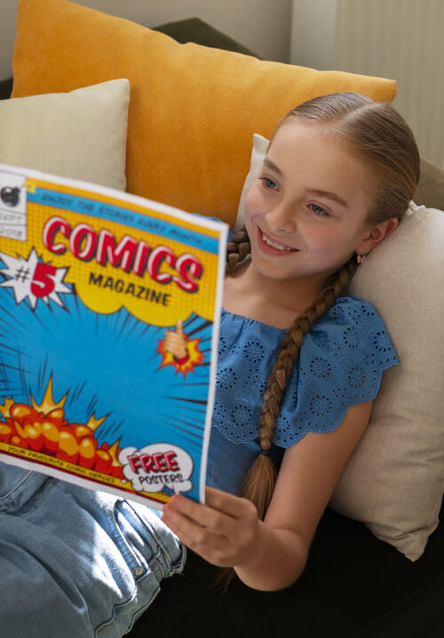 A imagem mostra uma menina sorridente, com longas tranças, lendo uma revista em quadrinhos enquanto está deitada em um sofá confortável. Ela usa uma blusa azul rendada e parece relaxada e imersa na leitura, com almofadas coloridas ao seu redor, criando um ambiente aconchegante e acolhedor.