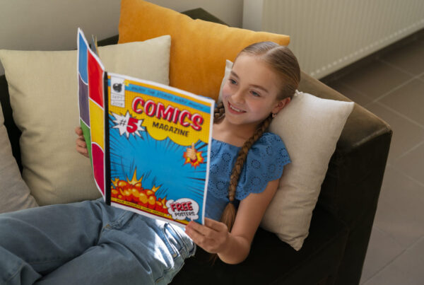 A imagem mostra uma menina sorridente, com longas tranças, lendo uma revista em quadrinhos enquanto está deitada em um sofá confortável. Ela usa uma blusa azul rendada e parece relaxada e imersa na leitura, com almofadas coloridas ao seu redor, criando um ambiente aconchegante e acolhedor.