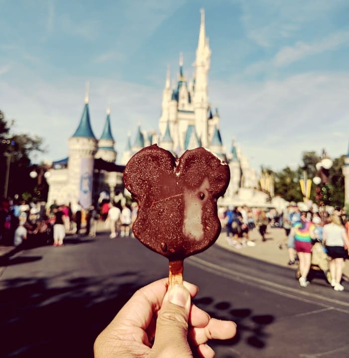 Um picolé coberto de chocolate, moldado no formato da cabeça de um famoso personagem de orelhas arredondadas, é segurado por uma mão em primeiro plano. Ao fundo, está o icônico castelo de conto de fadas com torres azuis, cercado por visitantes em um parque temático. O dia está ensolarado e o céu azul complementa a atmosfera mágica da cena.