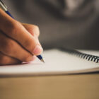 Pessoa escrevendo com caneta azul em caderno de espiral, representando o ato de praticar a escrita manual ou treinar habilidades de escrita.