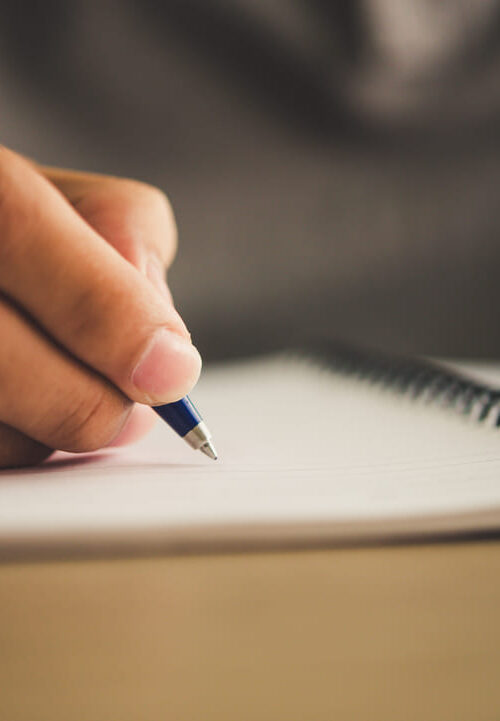 Pessoa escrevendo com caneta azul em caderno de espiral, representando o ato de praticar a escrita manual ou treinar habilidades de escrita.