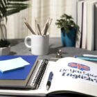 Uma mesa de estudos organizada com diversos materiais, incluindo um caderno aberto com a frase "Do you speak English?" acompanhada de uma bandeira do Reino Unido. Há também um livro azul com notas adesivas, lápis em uma caneca branca e pequenos vasos de plantas decorativos. Ao fundo, várias pilhas de livros estão dispostas.