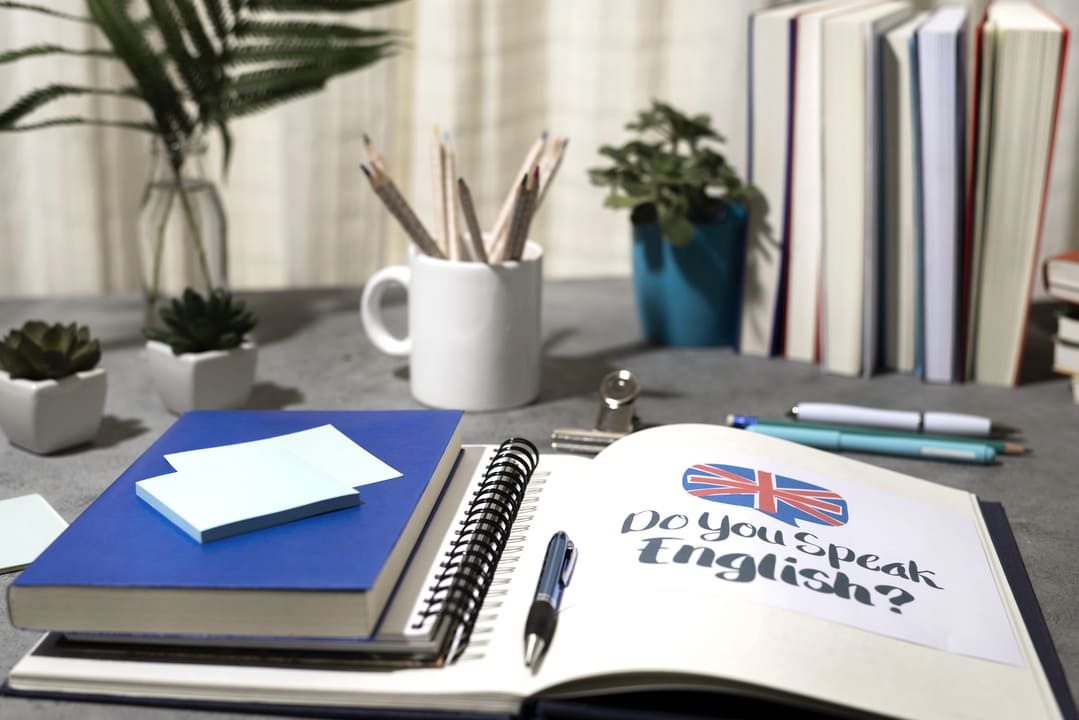 Uma mesa de estudos organizada com diversos materiais, incluindo um caderno aberto com a frase "Do you speak English?" acompanhada de uma bandeira do Reino Unido. Há também um livro azul com notas adesivas, lápis em uma caneca branca e pequenos vasos de plantas decorativos. Ao fundo, várias pilhas de livros estão dispostas.