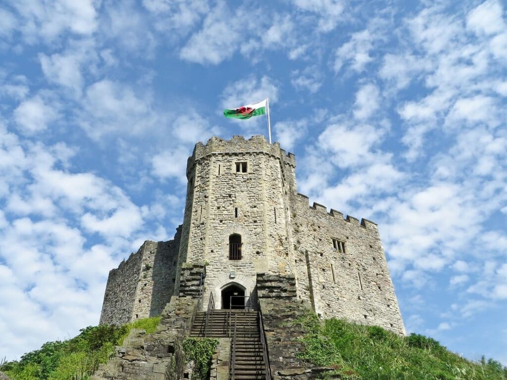 Uma antiga torre de castelo de pedra no topo de uma colina, com escadas de pedra levando à entrada principal. No topo da torre, uma bandeira do País de Gales está hasteada. O céu ao fundo está claro, com muitas nuvens brancas espalhadas.