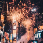 A imagem captura um espetáculo de fogos de artifício iluminando o céu em uma grande cidade durante a noite. Há arranha-céus iluminados ao fundo com painéis digitais e anúncios brilhantes, sugerindo um local movimentado e icônico como Times Square. No primeiro plano, silhuetas de pessoas observam e registram o momento com seus celulares. A cena transmite a energia de uma celebração, como o Réveillon, com um contraste marcante entre os fogos dourados e o céu escuro.