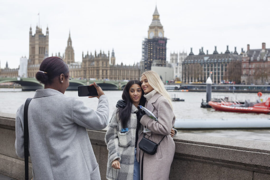 Três mulheres estão interagindo em um cenário icônico de Londres. Uma delas está tirando uma foto das outras duas, que estão abraçadas e posando sorridentes. Ao fundo, é possível ver o famoso Parlamento Britânico, o Big Ben (em reforma) e a ponte de Westminster sobre o rio Tâmisa. O dia está nublado, e a atmosfera é típica de Londres.