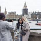 Três mulheres estão interagindo em um cenário icônico de Londres. Uma delas está tirando uma foto das outras duas, que estão abraçadas e posando sorridentes. Ao fundo, é possível ver o famoso Parlamento Britânico, o Big Ben (em reforma) e a ponte de Westminster sobre o rio Tâmisa. O dia está nublado, e a atmosfera é típica de Londres.