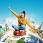 Uma jovem vestindo um moletom amarelo e calça jeans está sentada sobre uma mala vermelha, flutuando sobre um globo terrestre. Ela tem uma expressão alegre, com um braço estendido como se estivesse surfando no ar. Ao fundo, estão representados marcos turísticos icônicos, como a Estátua da Liberdade, o Big Ben e a Torre Eiffel, com um avião voando no céu azul.