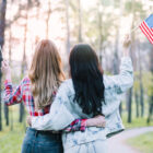 Duas mulheres estão abraçadas de costas para a câmera em um parque arborizado, segurando pequenas bandeiras dos Estados Unidos. A mulher à esquerda tem cabelos longos e loiros e veste uma camisa xadrez vermelha. A mulher à direita tem cabelos longos e escuros e veste um casaco jeans claro. O cenário tem árvores altas e um caminho sinuoso, com a luz suave do sol ao fundo.