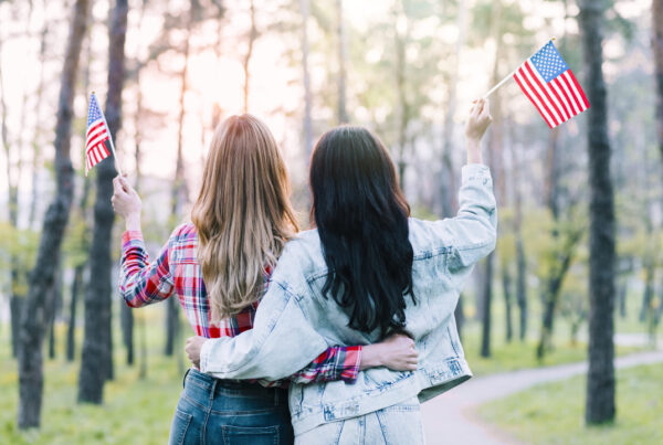 Duas mulheres estão abraçadas de costas para a câmera em um parque arborizado, segurando pequenas bandeiras dos Estados Unidos. A mulher à esquerda tem cabelos longos e loiros e veste uma camisa xadrez vermelha. A mulher à direita tem cabelos longos e escuros e veste um casaco jeans claro. O cenário tem árvores altas e um caminho sinuoso, com a luz suave do sol ao fundo.