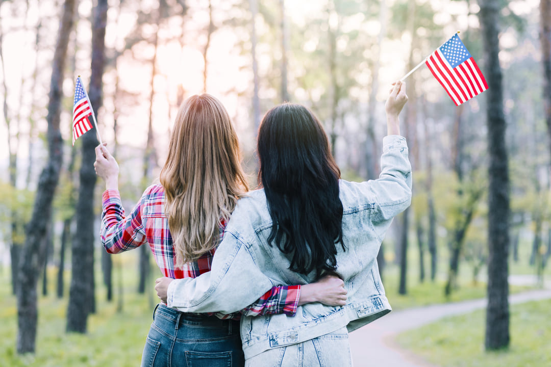 Duas mulheres estão abraçadas de costas para a câmera em um parque arborizado, segurando pequenas bandeiras dos Estados Unidos. A mulher à esquerda tem cabelos longos e loiros e veste uma camisa xadrez vermelha. A mulher à direita tem cabelos longos e escuros e veste um casaco jeans claro. O cenário tem árvores altas e um caminho sinuoso, com a luz suave do sol ao fundo.