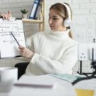 Professora de inglês ensinando tempos verbais em uma aula online, segurando um quadro branco enquanto grava com um celular apoiado em um tripé.