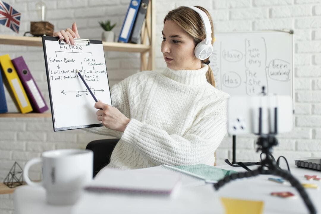 Professora de inglês ensinando tempos verbais em uma aula online, segurando um quadro branco enquanto grava com um celular apoiado em um tripé.