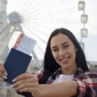 Mulher jovem sorrindo segura um passaporte azul e um cartão de embarque em frente a uma roda-gigante ao ar livre, aparentando felicidade e expectativa por uma viagem.