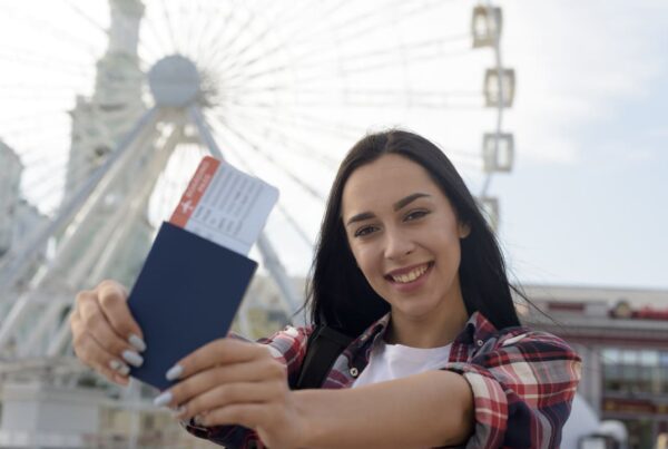 Mulher jovem sorrindo segura um passaporte azul e um cartão de embarque em frente a uma roda-gigante ao ar livre, aparentando felicidade e expectativa por uma viagem.