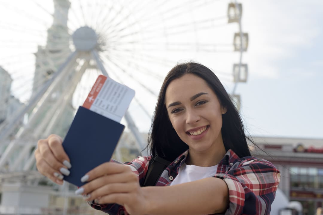 Mulher jovem sorrindo segura um passaporte azul e um cartão de embarque em frente a uma roda-gigante ao ar livre, aparentando felicidade e expectativa por uma viagem.