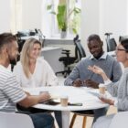 Quatro profissionais estão reunidos ao redor de uma mesa branca em um ambiente de escritório moderno e iluminado. Eles parecem estar em uma conversa descontraída, com expressões sorridentes. No fundo, há mesas com computadores, cadeiras ergonômicas e plantas decorativas, criando um ambiente de trabalho colaborativo.