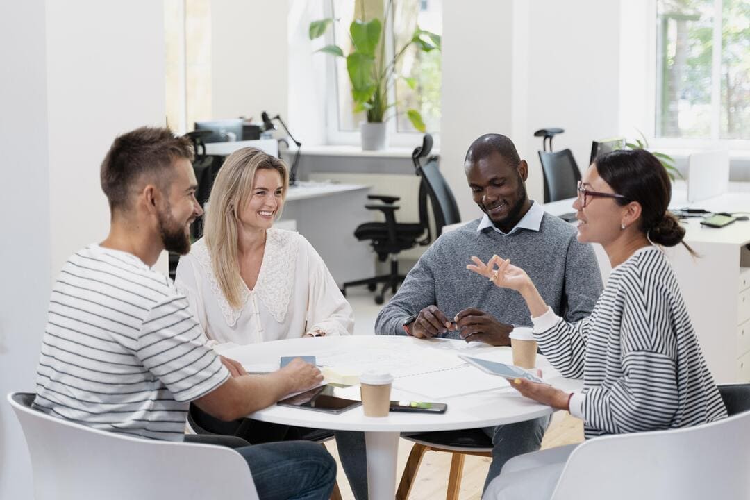 Quatro profissionais estão reunidos ao redor de uma mesa branca em um ambiente de escritório moderno e iluminado. Eles parecem estar em uma conversa descontraída, com expressões sorridentes. No fundo, há mesas com computadores, cadeiras ergonômicas e plantas decorativas, criando um ambiente de trabalho colaborativo.