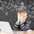 Imagem conceitual de uma criança loira, vestindo uma blusa de mangas listrada, cobrindo o rosto com as mãos em frente a um notebook branco. O fundo contém um quadro-negro digitalizado com letras e símbolos flutuando, representando dificuldades de aprendizado.