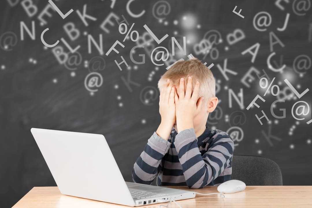 Imagem conceitual de uma criança loira, vestindo uma blusa de mangas listrada, cobrindo o rosto com as mãos em frente a um notebook branco. O fundo contém um quadro-negro digitalizado com letras e símbolos flutuando, representando dificuldades de aprendizado.