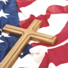 Uma cruz de madeira repousa sobre uma bandeira dos Estados Unidos, que está levemente amassada, com suas cores vibrantes de azul, vermelho e branco.