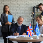Grupo de executivos assinando documentos em uma mesa com as bandeiras do Reino Unido e da União Europeia à frente, representando uma negociação ou acordo internacional.
