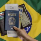 Pessoa segurando um passaporte brasileiro e uma carteira com cartões, passagens e documentos, com a bandeira do Brasil ao fundo.