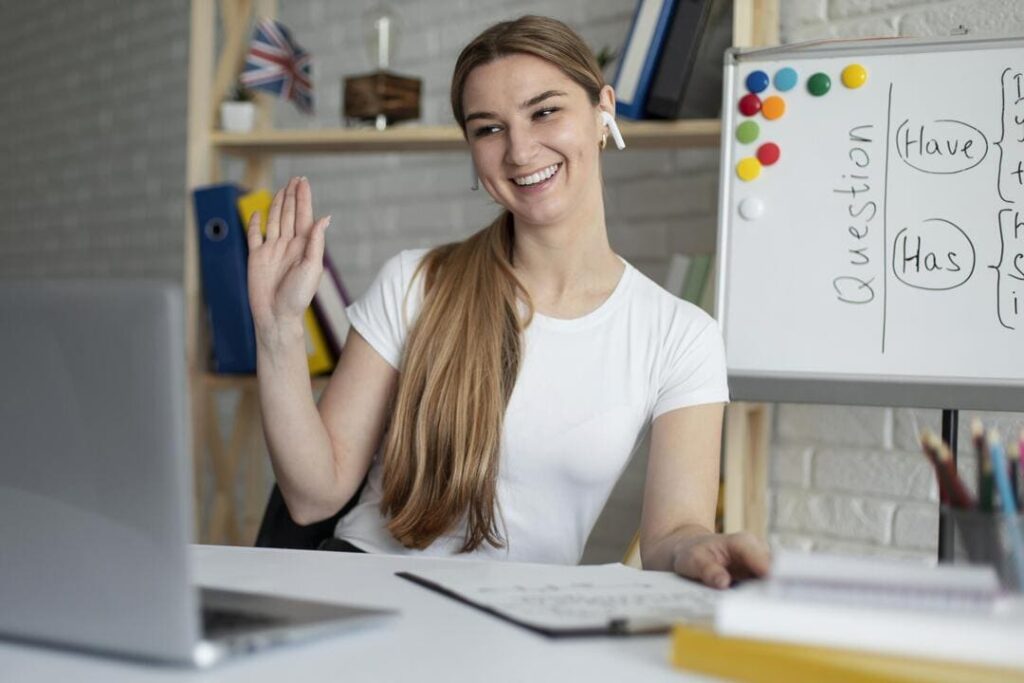 Mulher sorridente acena para o computador durante uma videochamada, com um quadro branco ao lado mostrando a estrutura gramatical "Have" e "Has".