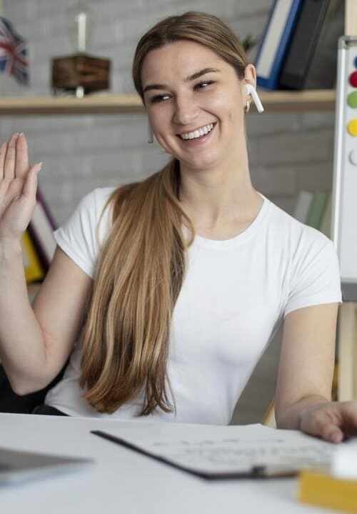 Mulher sorridente acena para o computador durante uma videochamada, com um quadro branco ao lado mostrando a estrutura gramatical "Have" e "Has".